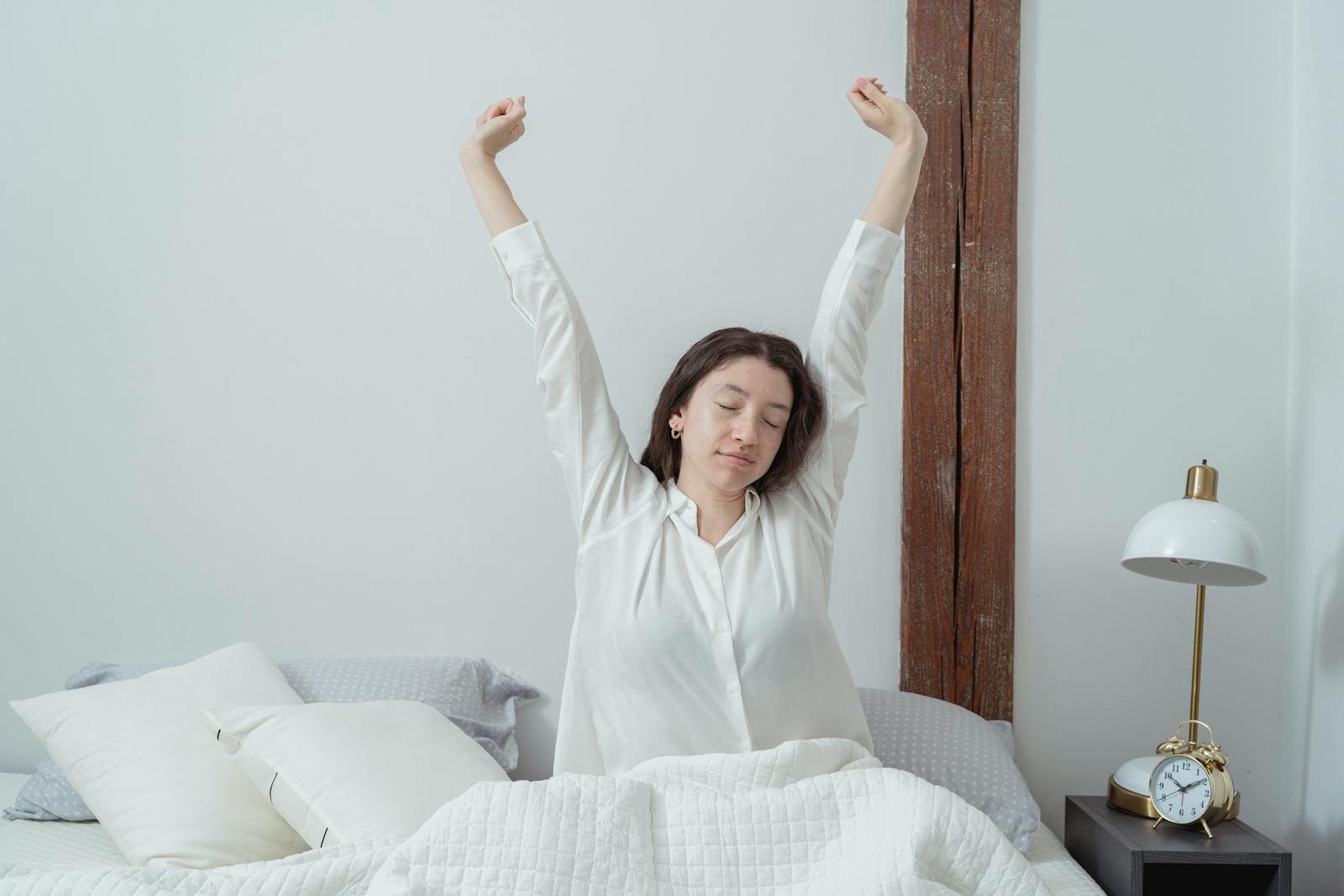 Adulto sentado na beira da cama com a mão no pescoço em expressão de dor matinal, em quarto iluminado por luz natural, cenário típico de torcicolo ao acordar.